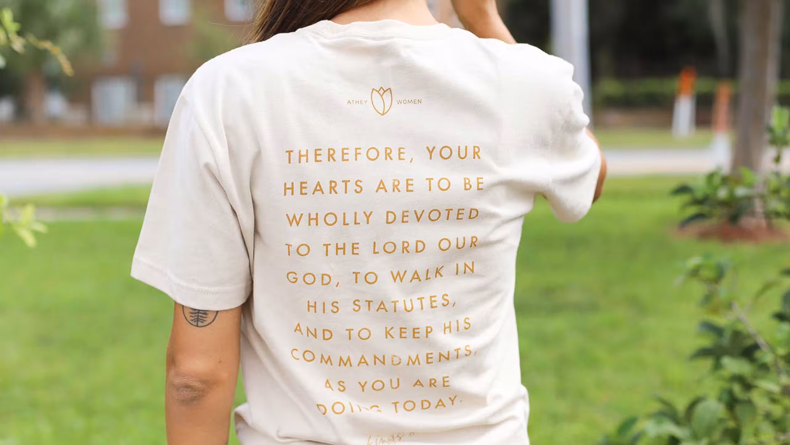 the back of a women wearing a cream white t-shirt with gold writing printed on it